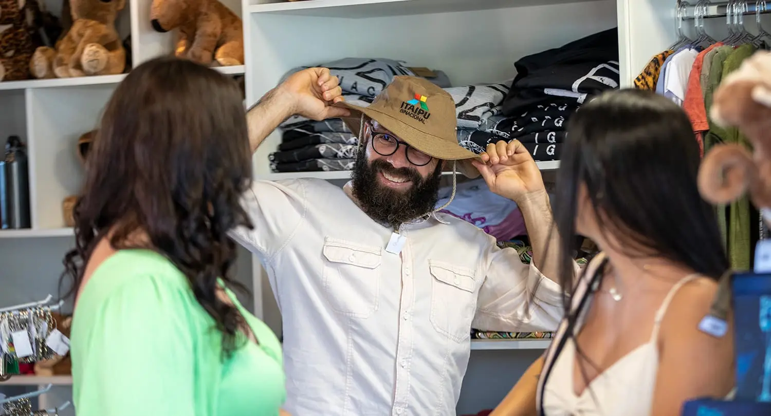 Homem usando chapéu com logo da Usina Hidrelétrica de Itaipu, ao lado de duas mulheres olhando para ele.