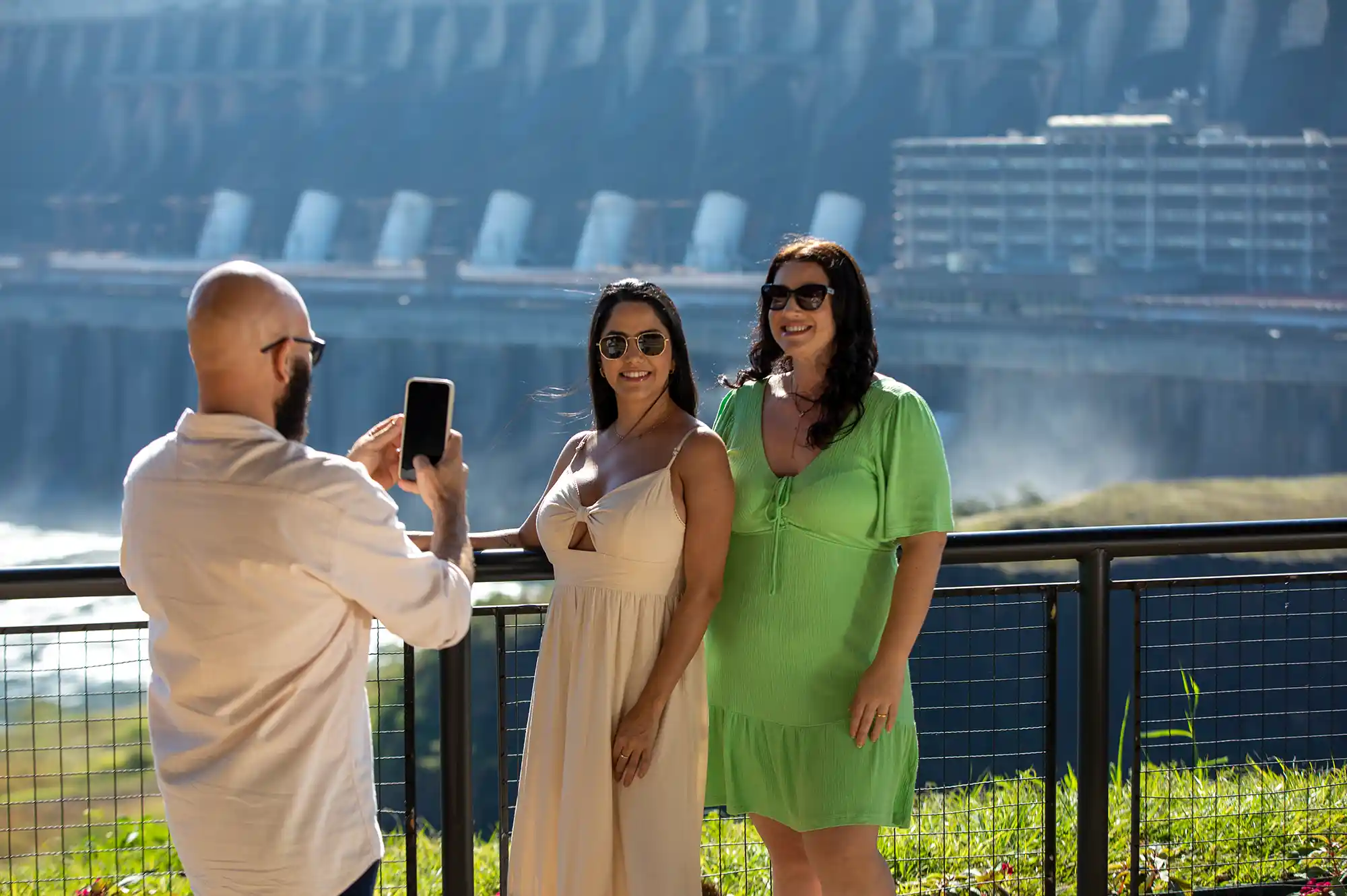 Duas mulheres posando para foto sendo tirada por homem com a Usina Hidrelétrica de Itaipu ao fundo.